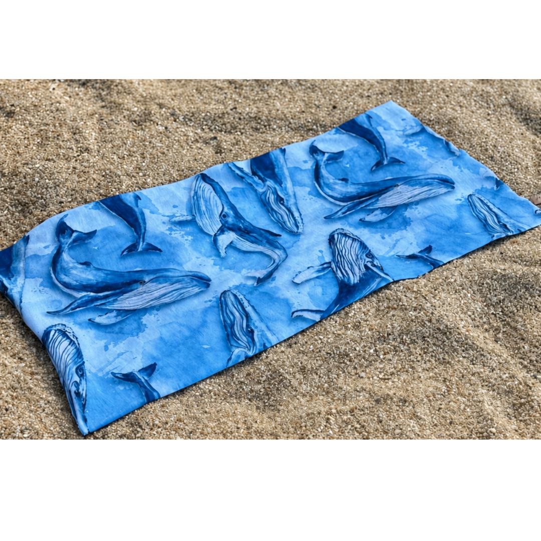 Humpback Whale Sun Buff with a blue ocean-inspired watercolor print, displayed flat on a sandy beach