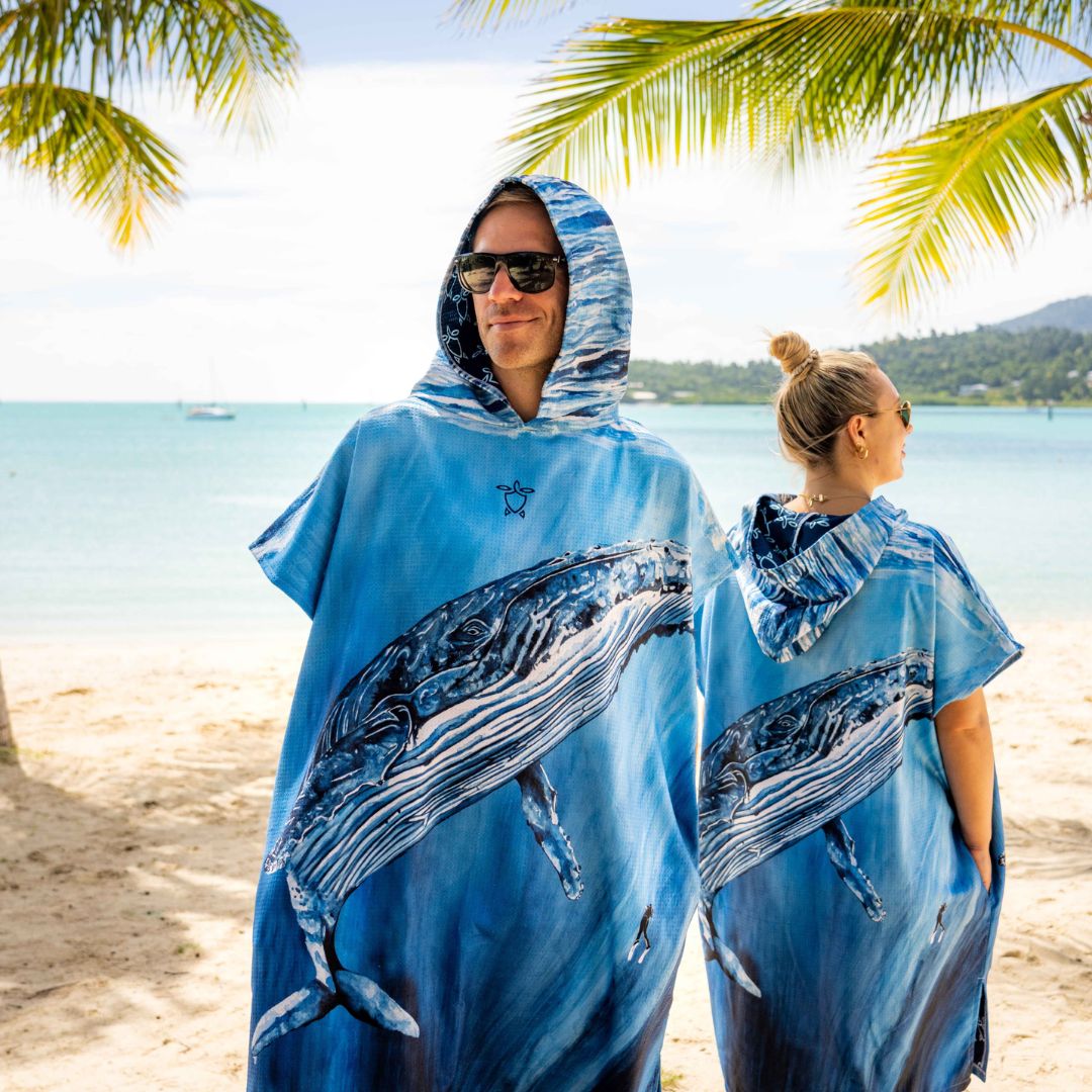 Two people wearing blue whale-print hooded ponchos standing on a sandy beach with the ocean and palm leaves in the background.