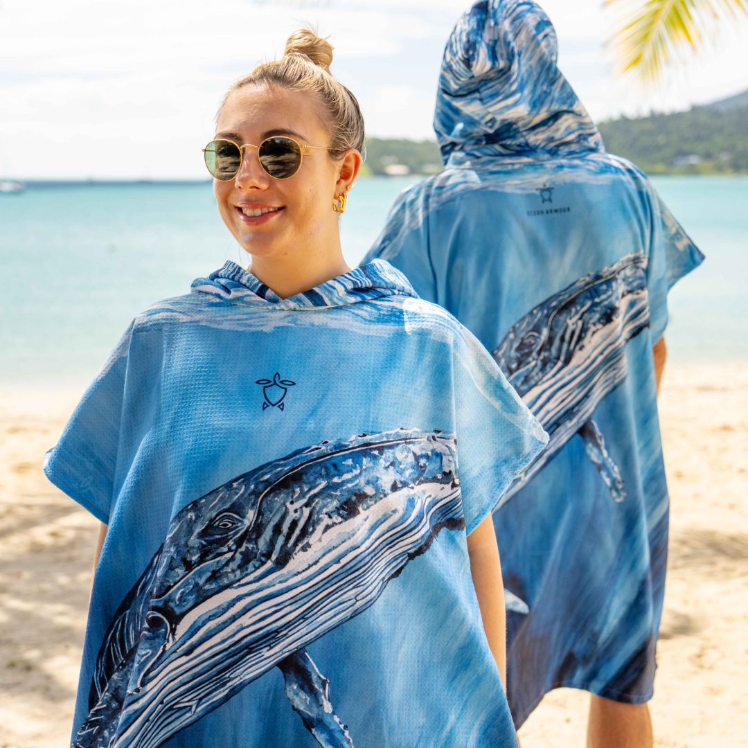 A woman on a beach wearing a blue whale-print hooded poncho, shown from both the front and back to display the design. The product features a large blue and white whale graphic with a small star logo on the chest.