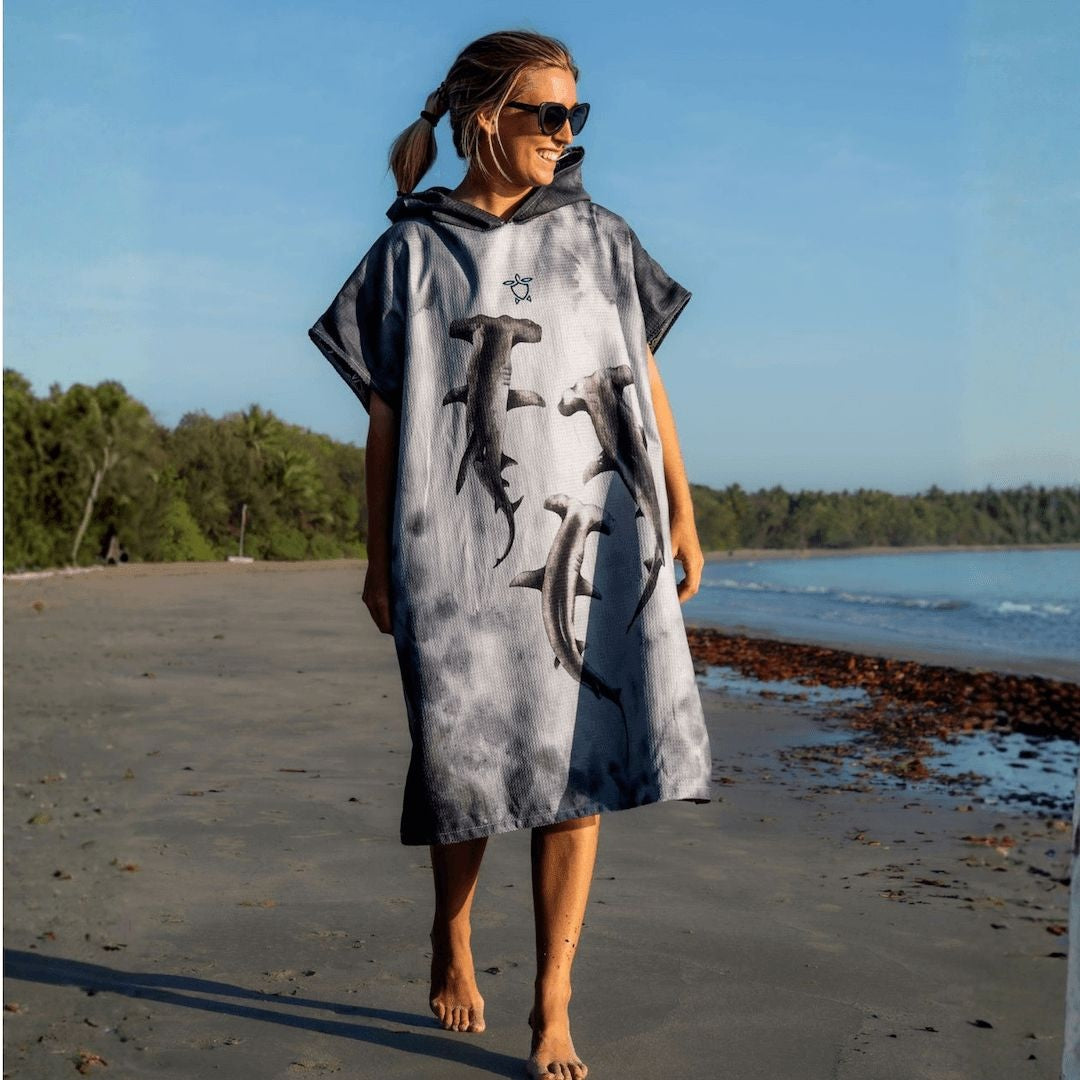 A woman wearing a gray and white tie-dye hooded poncho with shark prints walks barefoot on a sandy beach next to the ocean.