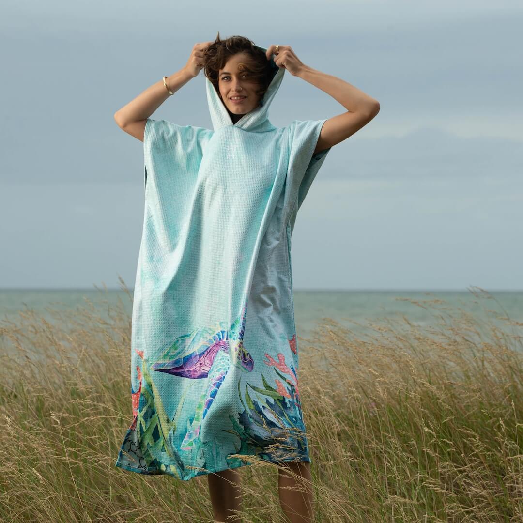 A woman wearing a light blue hooded beach towel poncho with a sea turtle design, standing in a grassy field with the ocean in the background.