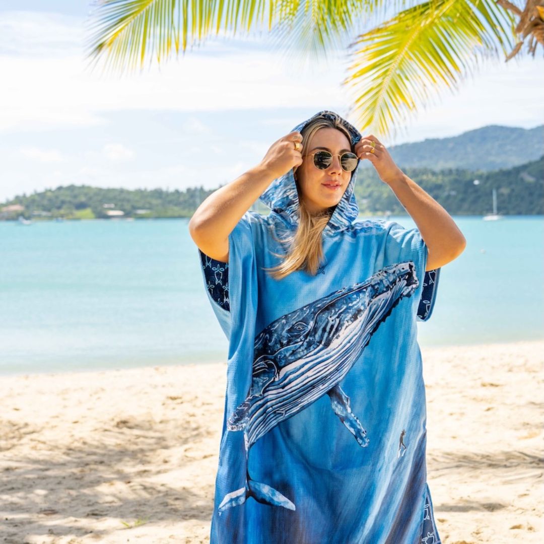 A woman wearing a blue whale-print beach cover-up and sunglasses stands on a sandy beach with the ocean and palm leaves in the background.