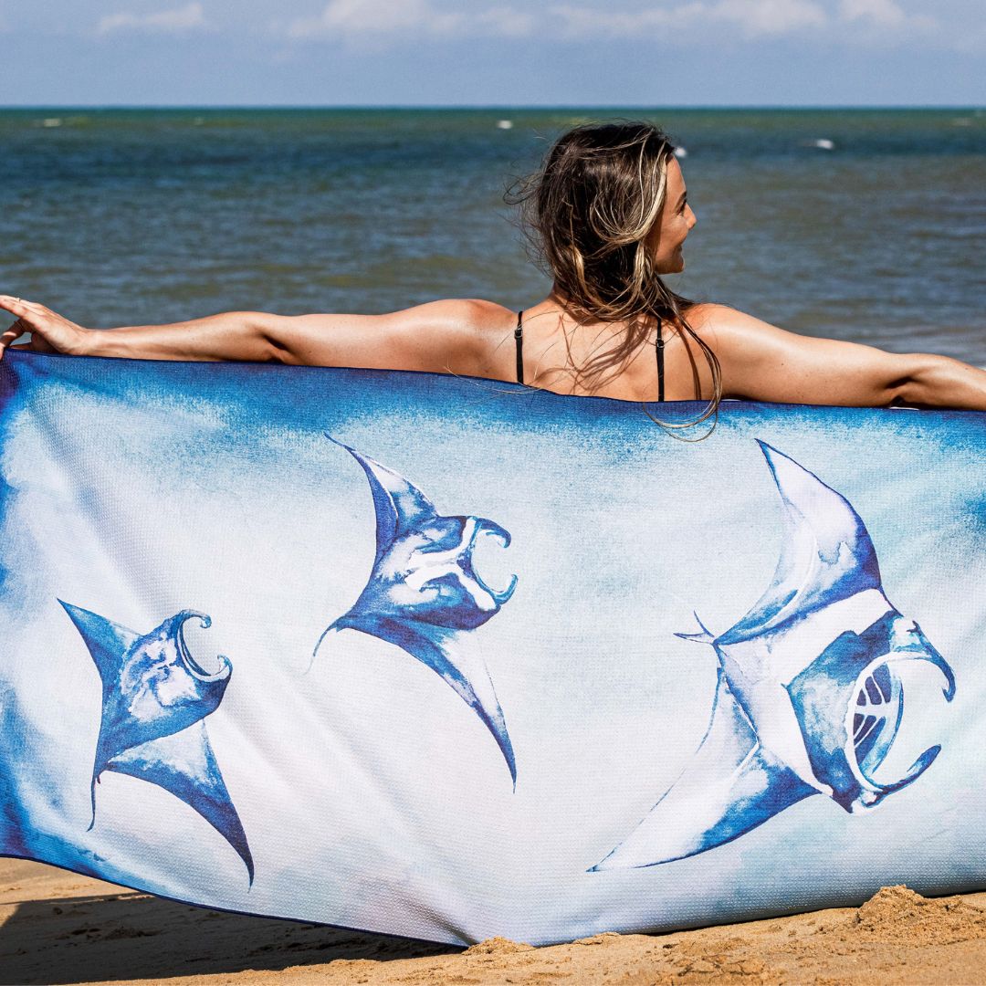 A woman on a beach holds up a large white and blue beach towel featuring illustrations of manta rays, with the ocean and sky in the background.