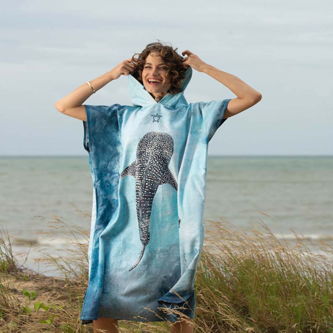 A woman stands on a beach dune wearing a light blue hooded towel poncho with a large whale shark graphic on the front. The background shows the ocean and cloudy sky.