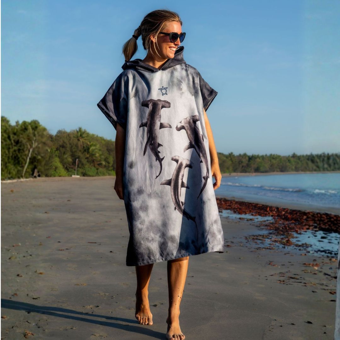 Woman walking barefoot along the beach wearing a hooded Hammerhead Shark Changing Poncho Towel featuring three hammerhead shark illustrations on the front.