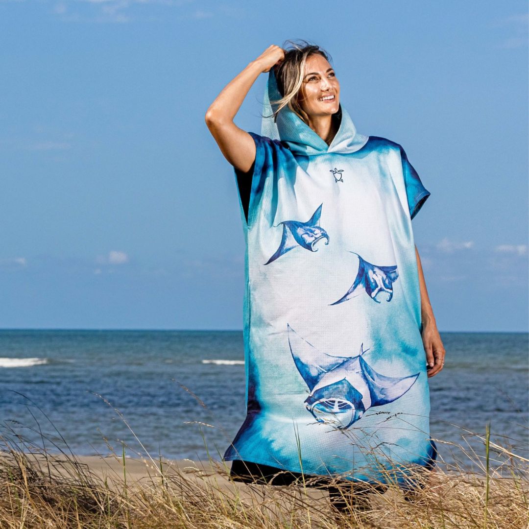 A woman standing on a beach dune wearing a hooded poncho-style cover-up with a blue and white manta ray print, with the ocean and blue sky in the background.