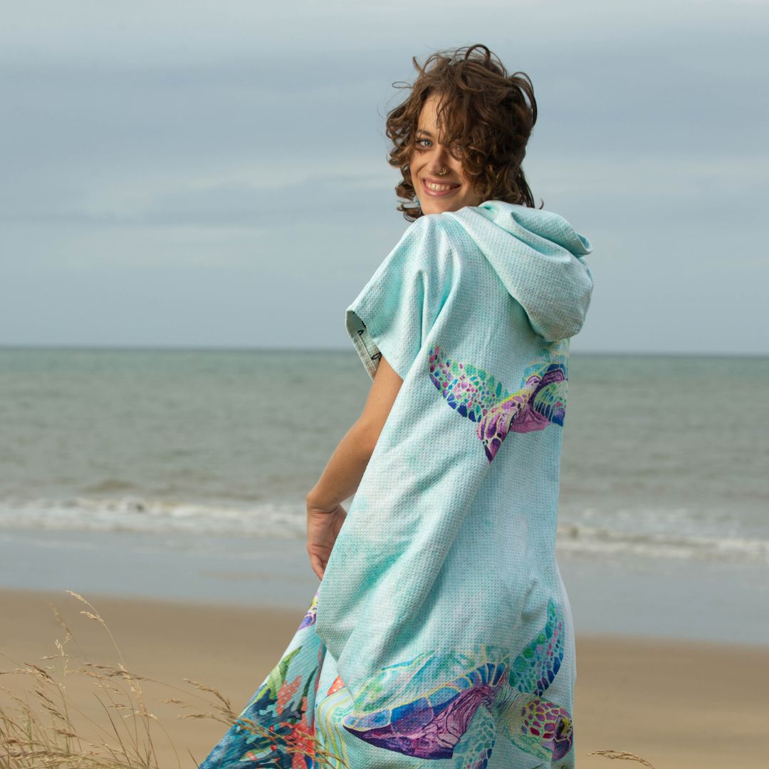 A woman standing on a beach dune wearing a light aqua hooded beach towel wrap with colorful turtle prints, looking back over her shoulder with a smile. The background shows the ocean and cloudy sky.