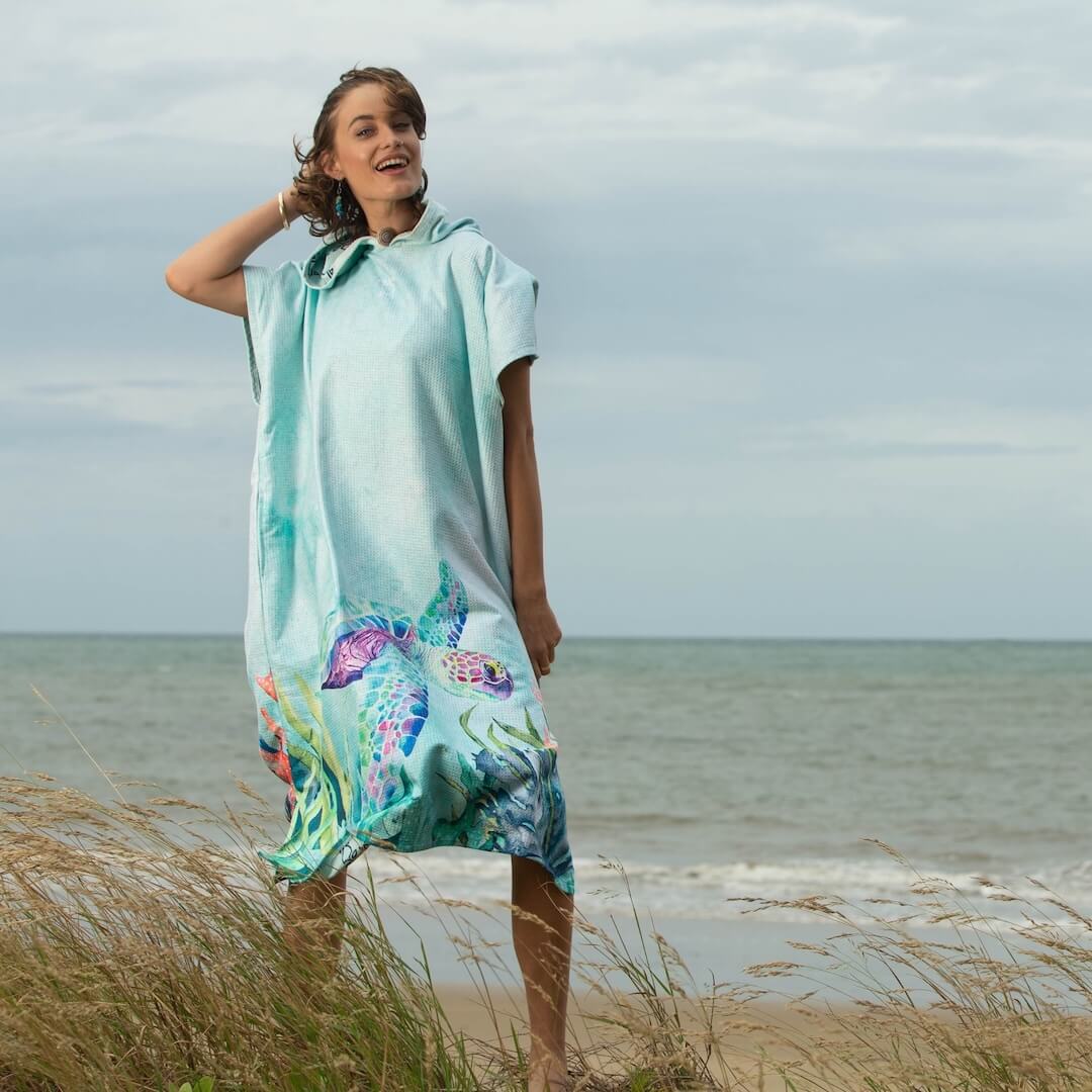 A woman stands on a beach dune wearing a light green hooded towel poncho with a water colour sea turtle print, with the ocean behind her.