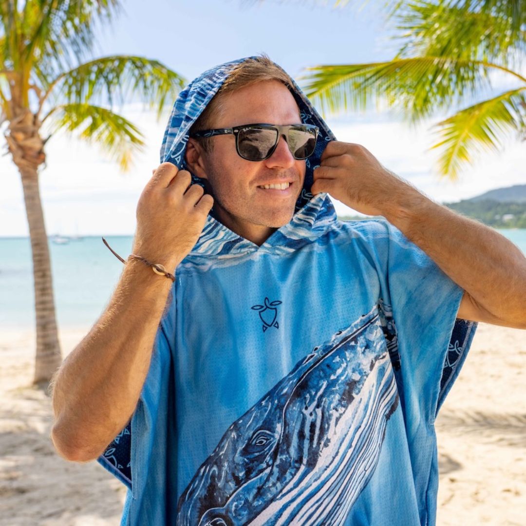 A man on a sunny beach wearing a blue hooded towel cover-up with a whale print and sunglasses, with palm trees and ocean in the background.