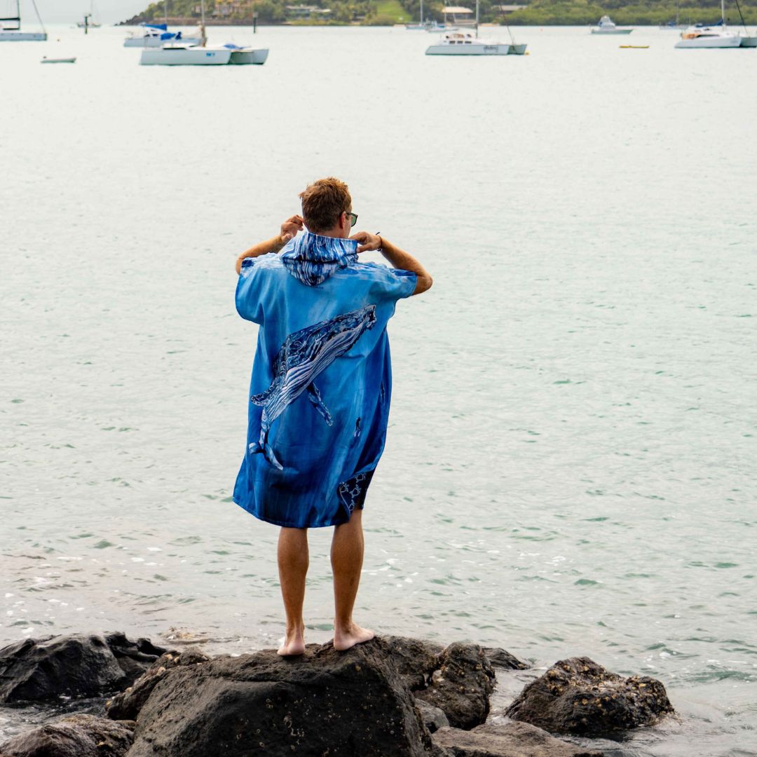 A man stands on dark rocks by the water, viewed from behind, wearing a blue hooded towel cover-up with a whale print. Boats are visible on the water in the background.