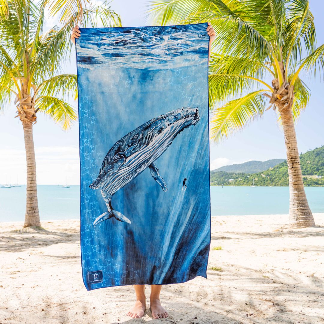 A person standing on a sandy beach holds up a large rectangular blue towel featuring a whale underwater print. Palm trees and a tropical ocean scene are visible in the background.