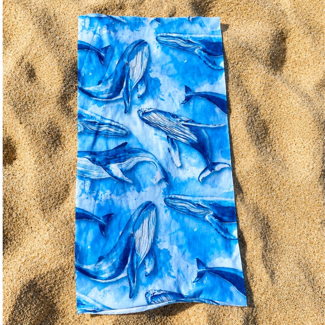 Blue Sun Buff with an all-over humpback whale print in a watercolor ocean design, shown flat on a sandy surface