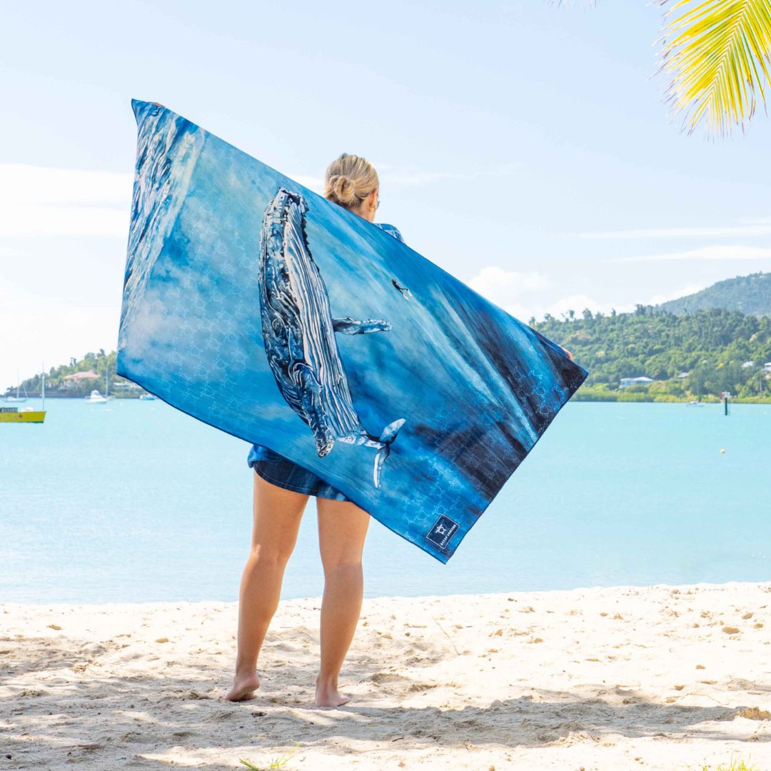 A woman on a sandy beach holds up a large blue towel featuring a whale print, with the ocean and distant hills in the background.
