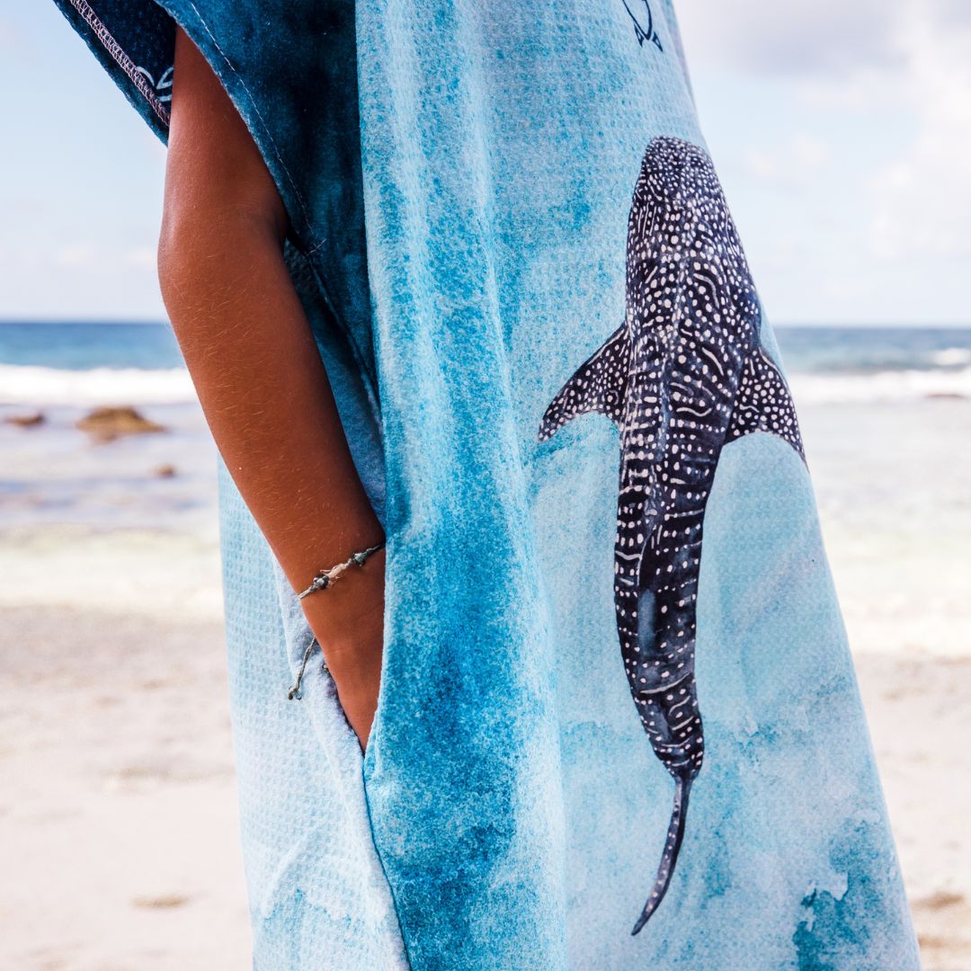 A person standing on a beach wearing a light blue towel or wrap with a large whale shark print and a dark blue hood, with the ocean and sand blurred in the background.