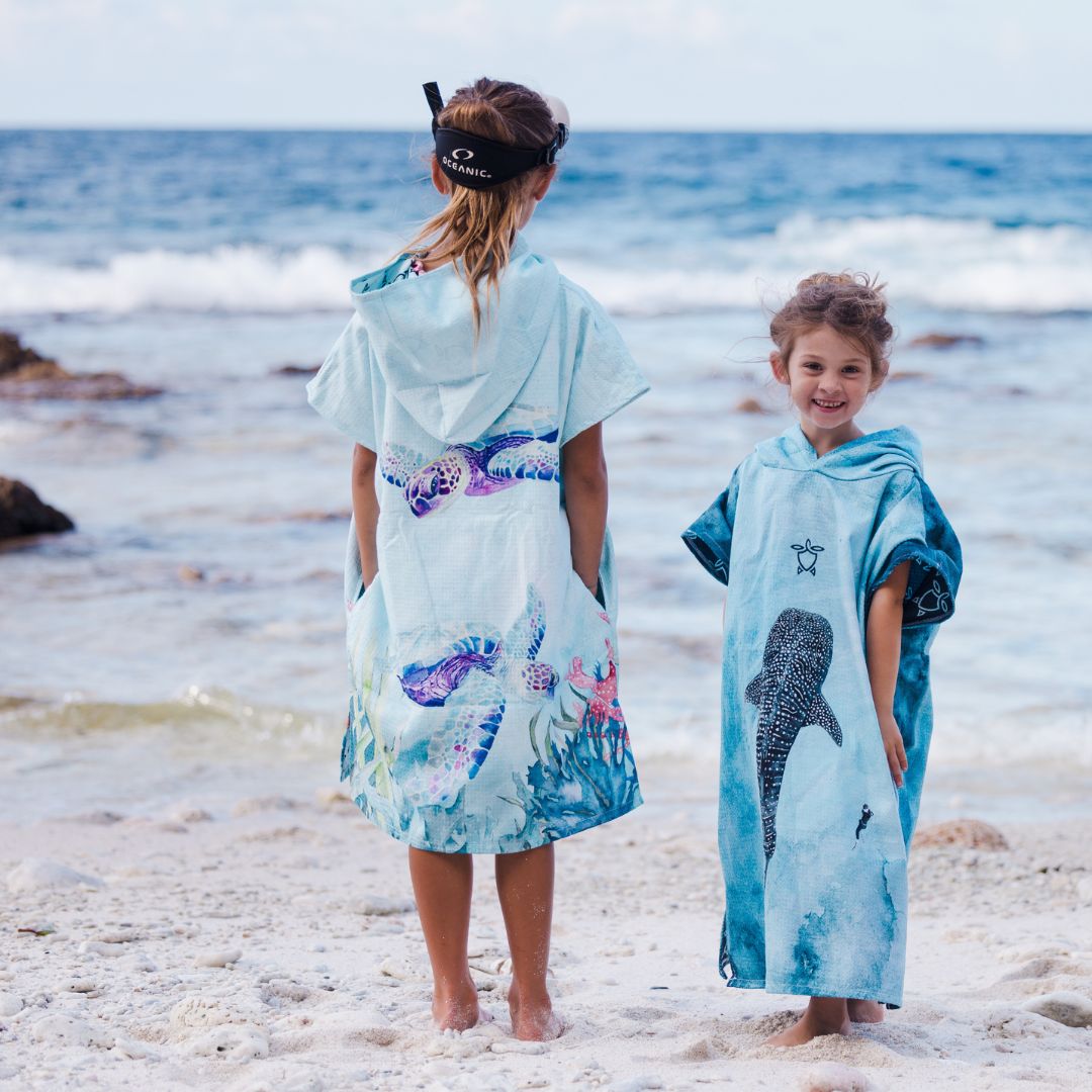 Two children standing on a sandy beach facing the ocean, wearing hooded towel ponchos with ocean-themed prints. The child on the left has a light blue towel poncho with purple and pink sea turtle designs, and the child on the right wears a blue towel poncho with a black spotted whale print and a Star of David.