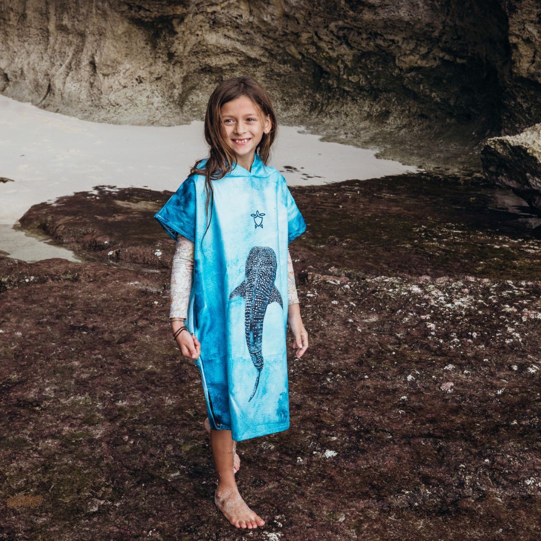 A young girl stands barefoot on a rocky beach wearing a bright blue poncho-style cover-up with a large shark graphic on the front.
