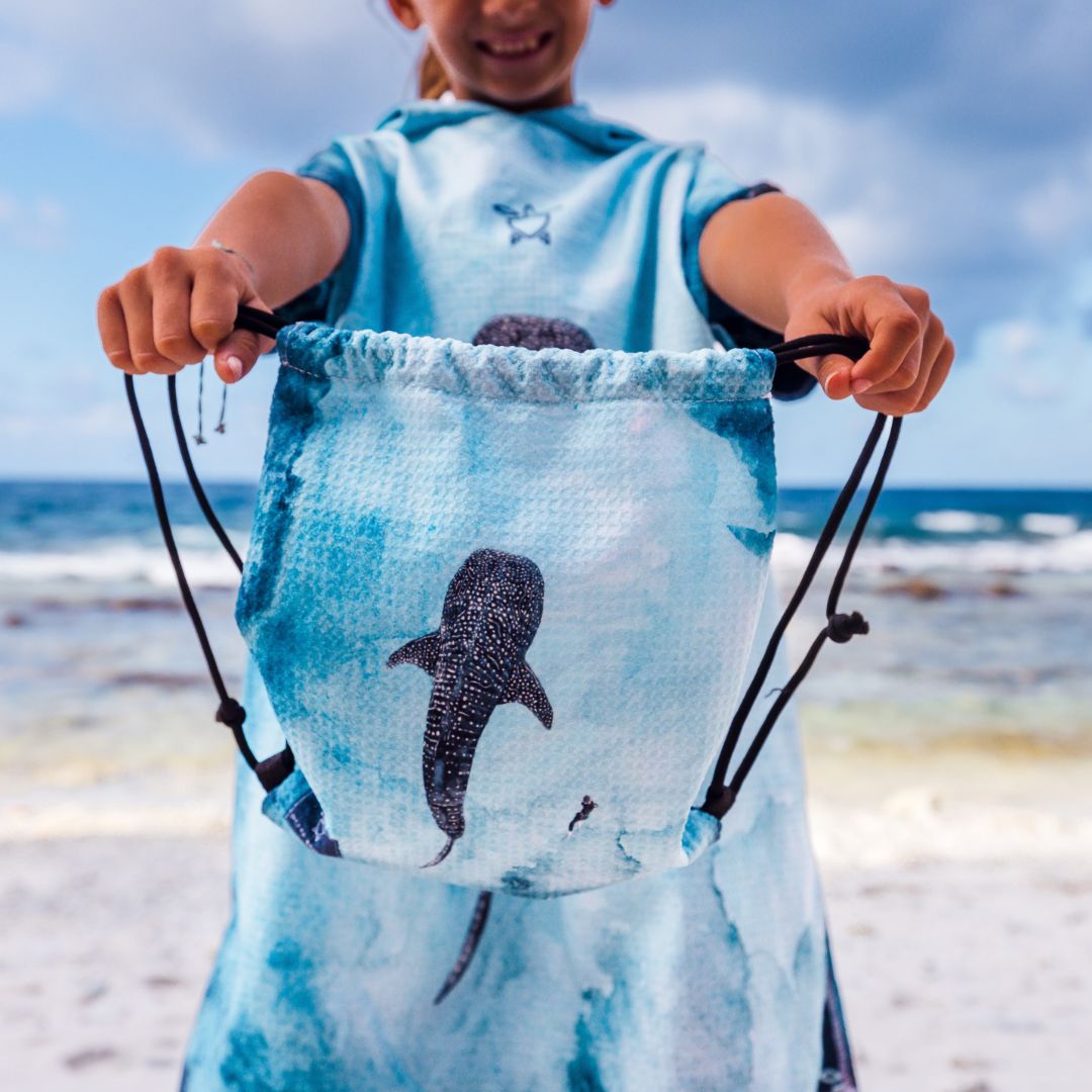 A child standing on a beach holding up a drawstring bag with a shark print. The bag is light blue with a black shark graphic and black drawstrings.