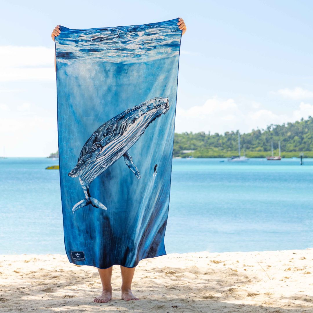 A person standing on a sandy beach holds up a large blue towel featuring a whale swimming underwater. The background shows a tropical ocean scene with boats and a shoreline.