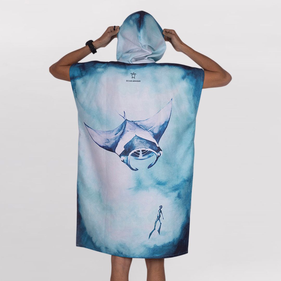 A person wearing a hooded beach towel or poncho with a watercolor-style manta ray and diver illustration in shades of blue and white, shown from the back against a light gray studio background.
