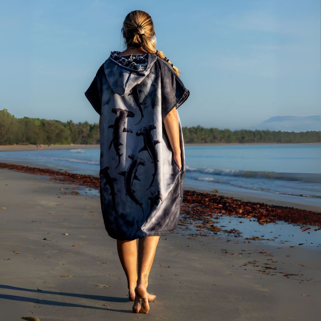 A woman stands on a beach facing the ocean, wearing a hooded towel or poncho with a shark print. The garment is light gray with black shark silhouettes and a dark patterned hood.