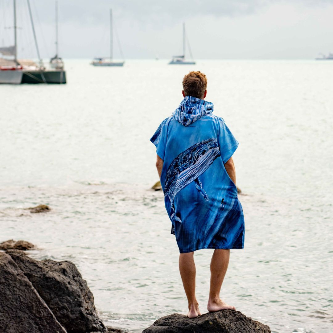 A man stands on a rocky beach facing the ocean, wearing a blue hooded towel or poncho with a large whale print. Sailboats are visible on the water under an overcast sky.