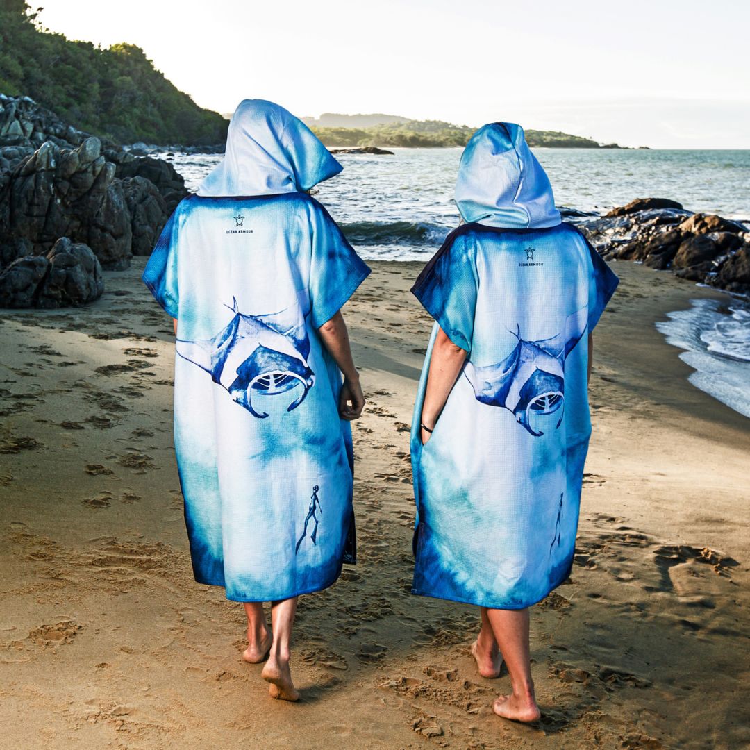 Two adults wearing hooded Manta Ray Changing Poncho Towels walk along the beach, showcasing the back design featuring a large blue manta ray illustration.