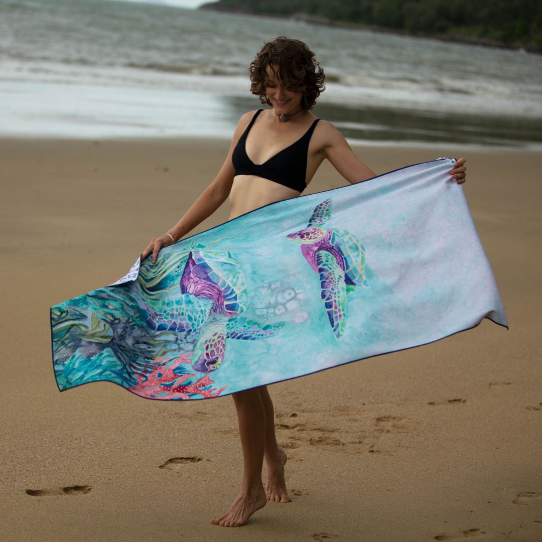 Female model displaying a large Ocean Armour microfiber beach towel with vibrant sea turtle illustrations and underwater reef artwork by the ocean.