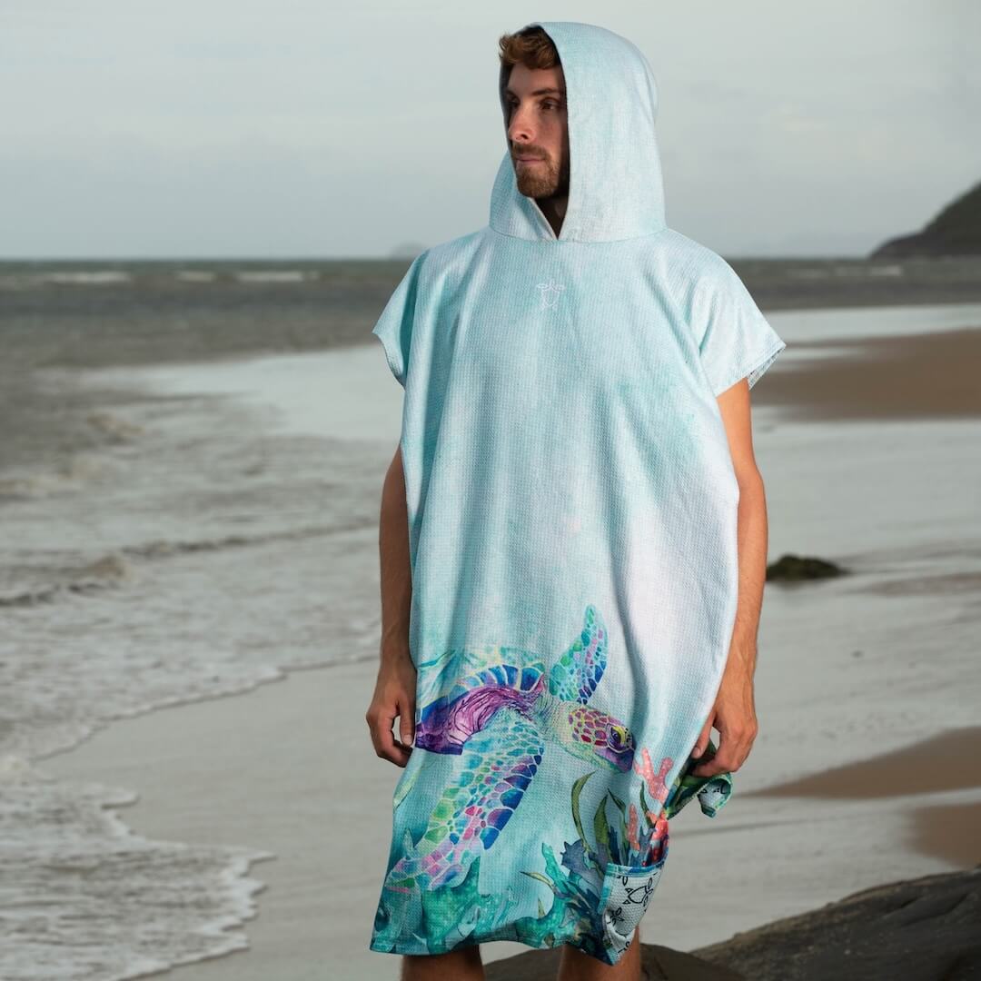 A man wearing a hooded beach poncho with a colorful sea turtle print, standing on a sandy beach.