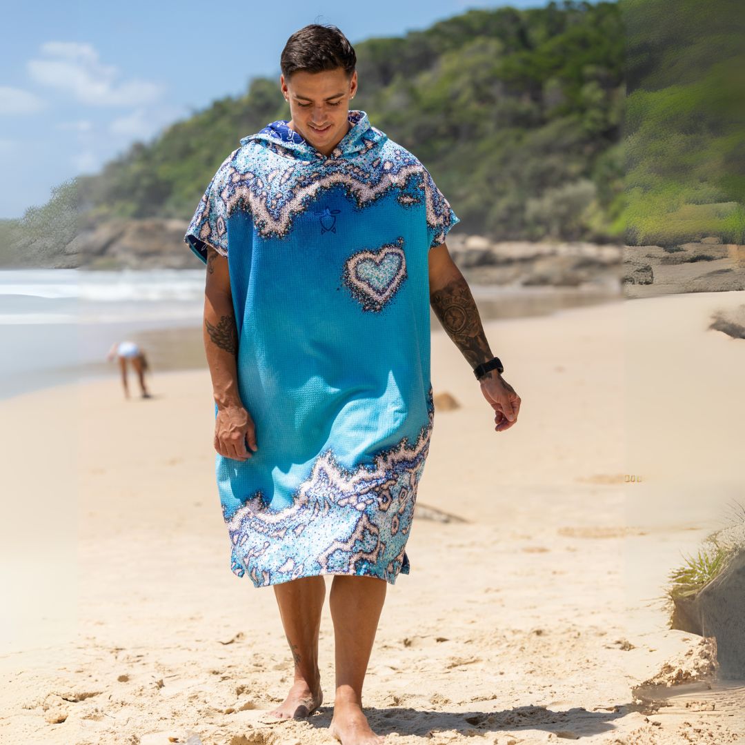A man walking on a sandy beach wearing a bright blue hooded kaftan-style cover-up with white and light blue paisley patterns and a heart-shaped patch on the chest.