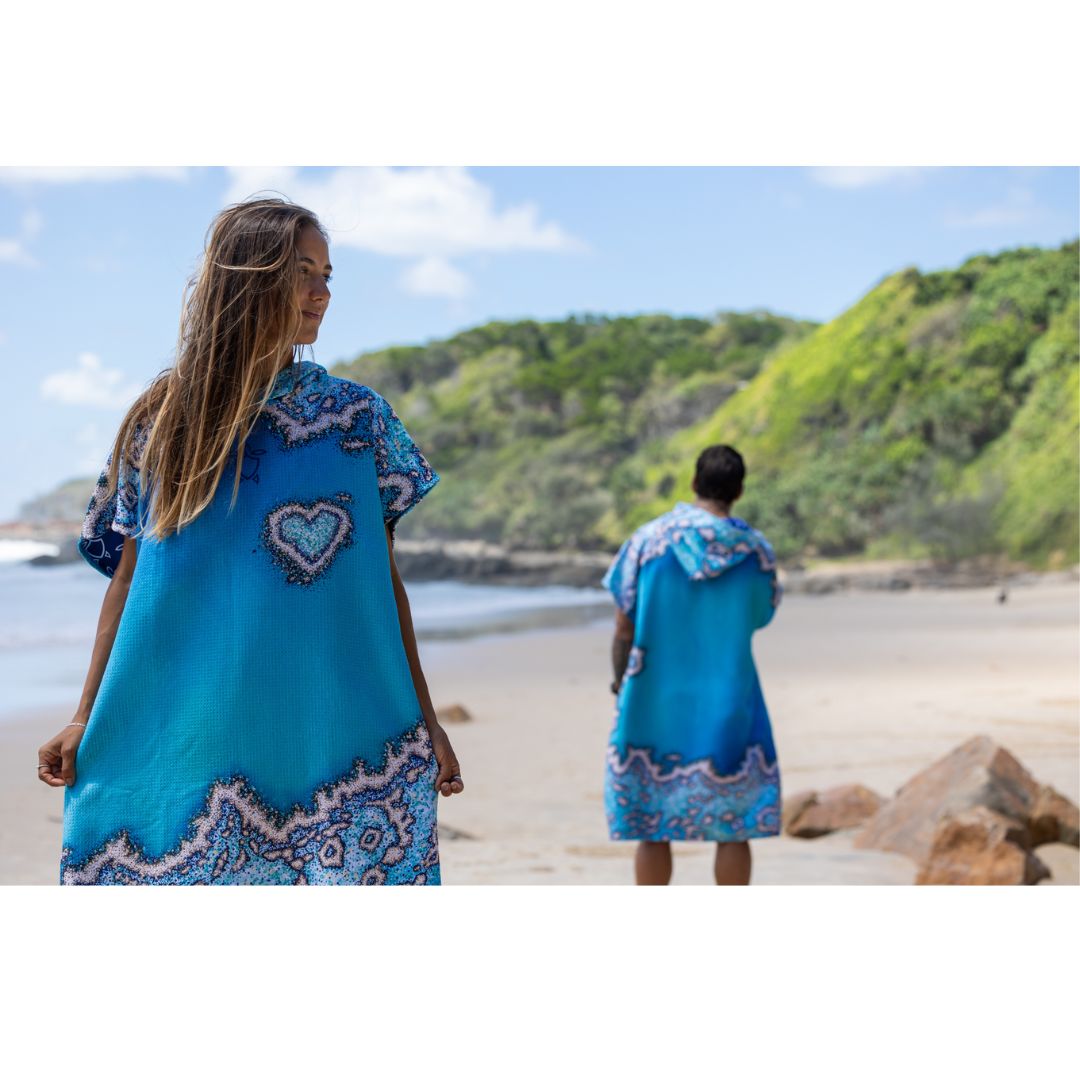 Two adults wearing Reef Love Hooded Changing Poncho Towels on a tropical beach, showcasing the front and back of the ocean-inspired design.