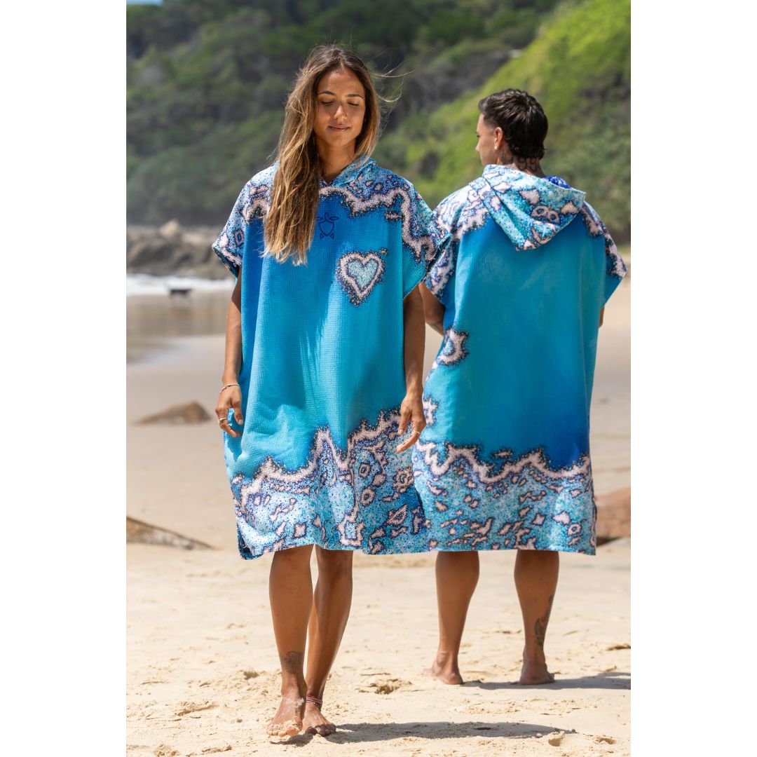 A man and a woman wearing matching bright blue beach ponchos with white and pink paisley-style borders and a heart motif, standing on a sandy beach with green hills in the background.
