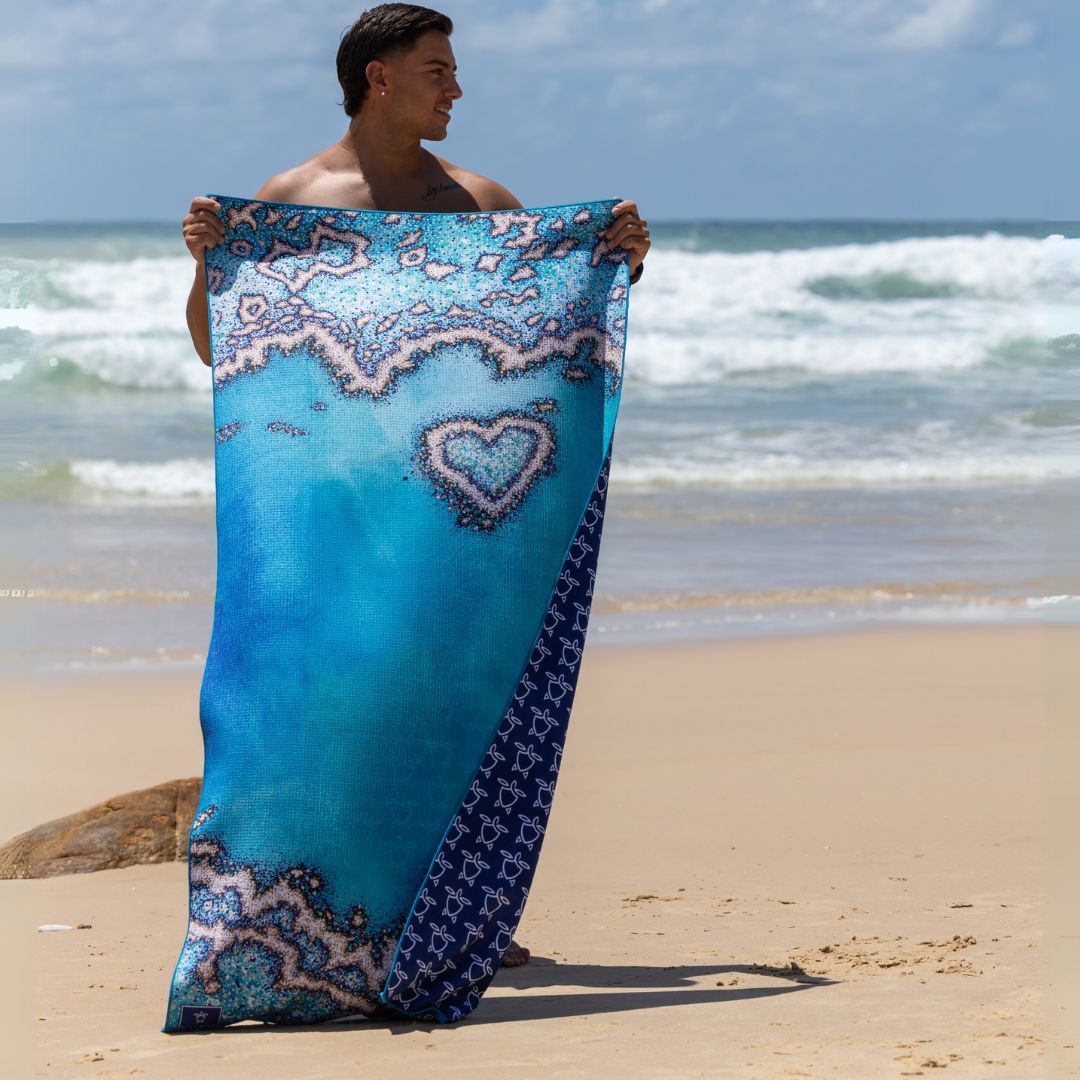 Person holding the Reef Love quick-dry sand-free beach towel on a sandy beach, featuring a blue ocean-inspired design with coral patterns and a heart-shaped reef motif