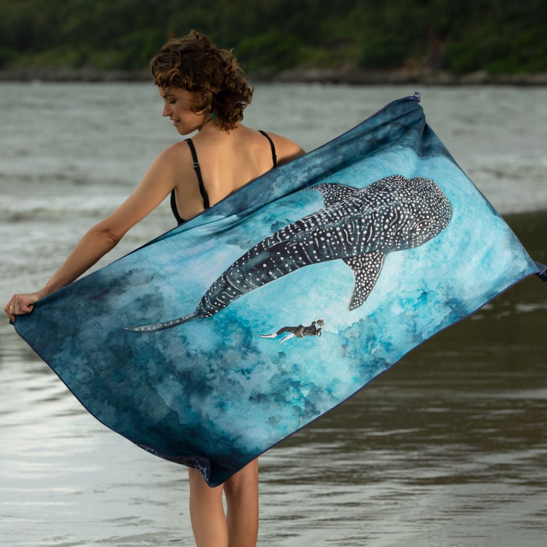 Woman holding an Ocean Armour Whale Shark sand-free beach towel on the shoreline. The quick-drying microfiber towel features a large whale shark swimming above a freediver against a blue watercolor ocean background, displayed across the full width of the towel.