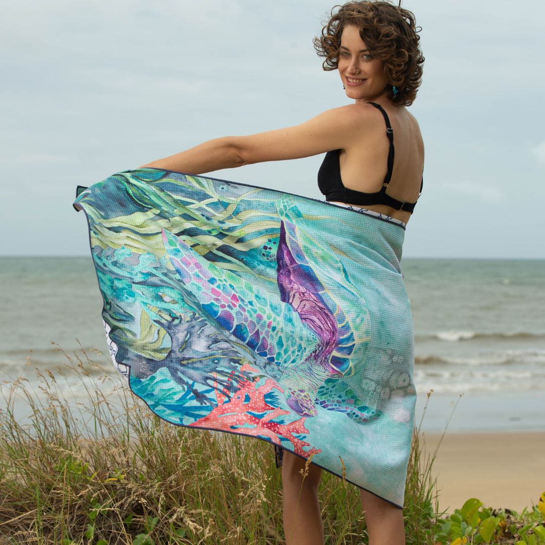 Ocean-inspired Sea Turtle beach towel with vibrant underwater artwork displayed by a woman against a scenic beach and rocky shoreline.