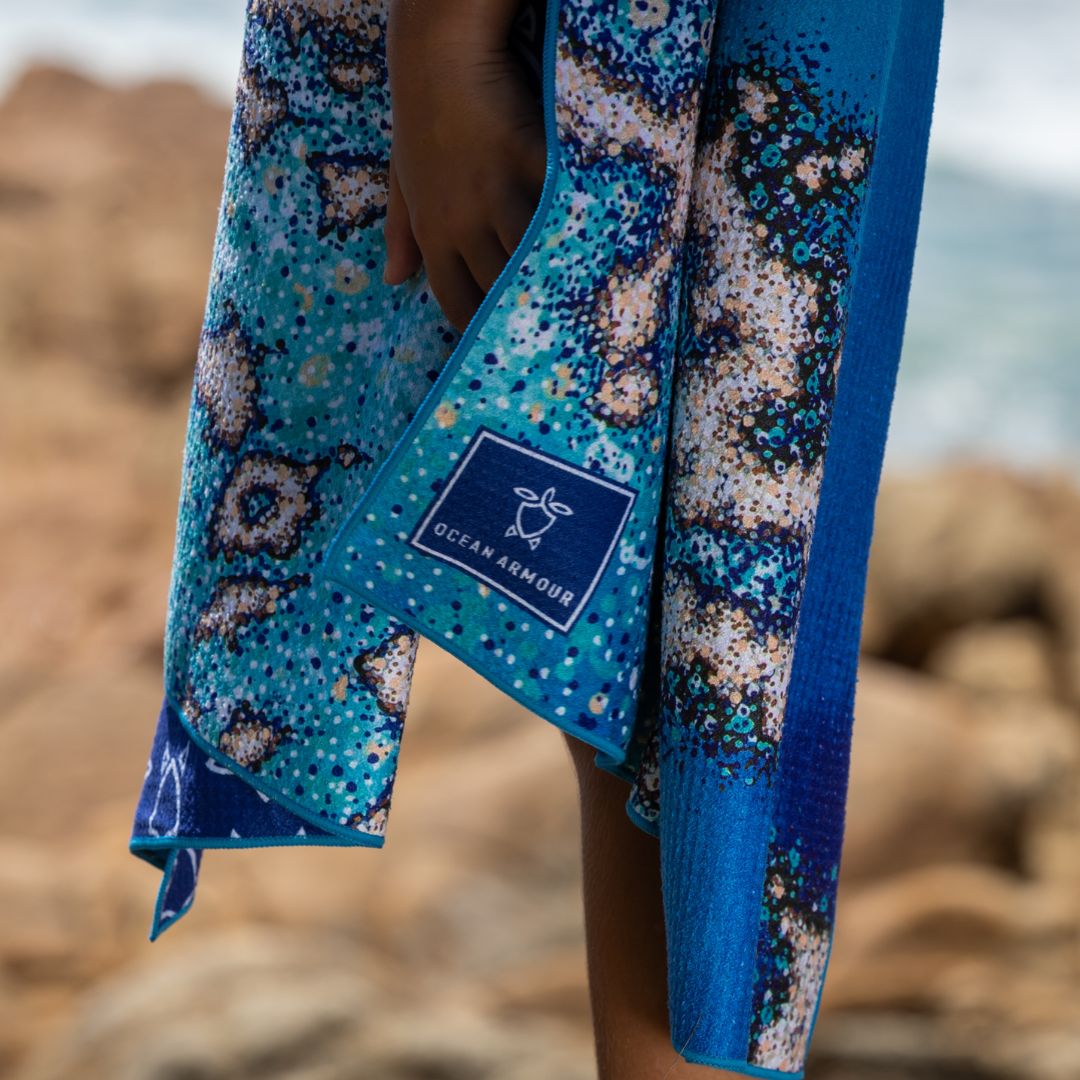 A close-up of a person wearing a blue patterned swim cover-up or sarong with a visible Ocean Armour label, photographed outdoors against a rocky beach background.