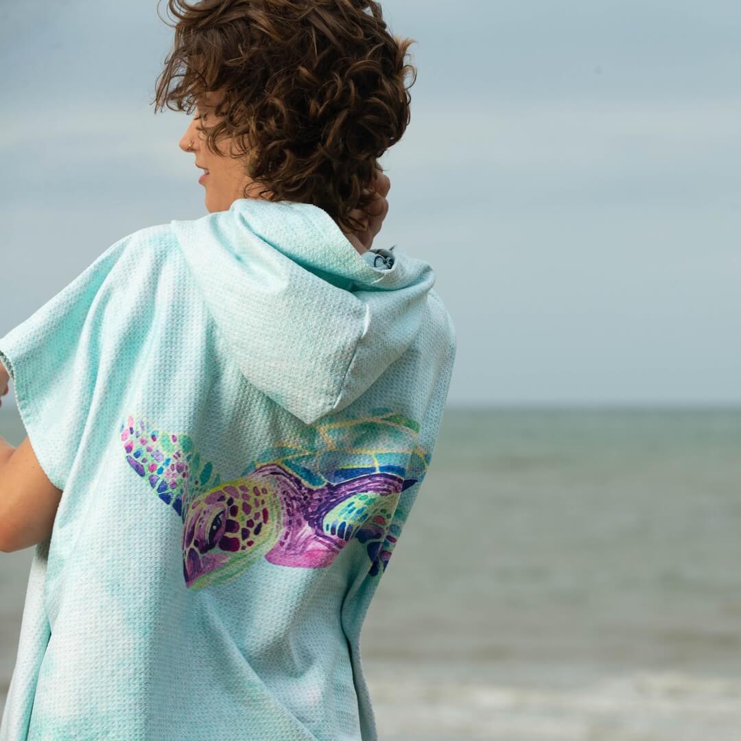 A woman standing outdoors by the ocean wearing a light aqua hooded towel poncho with a large watercolour sea turtle design on the back.