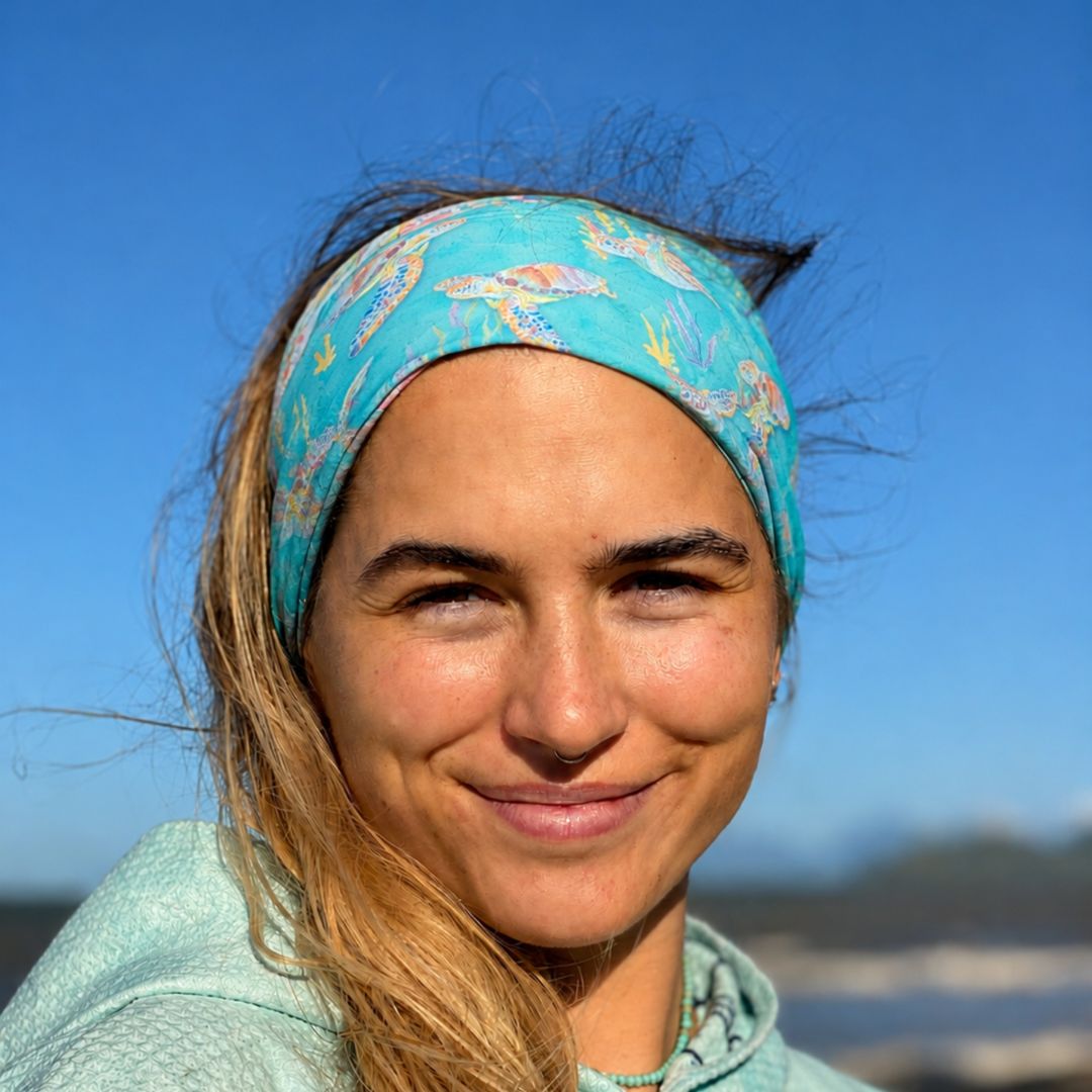 A smiling woman outdoors wearing a turquoise patterned headband and a light mint green top, with a blue sky and blurred shoreline in the background.