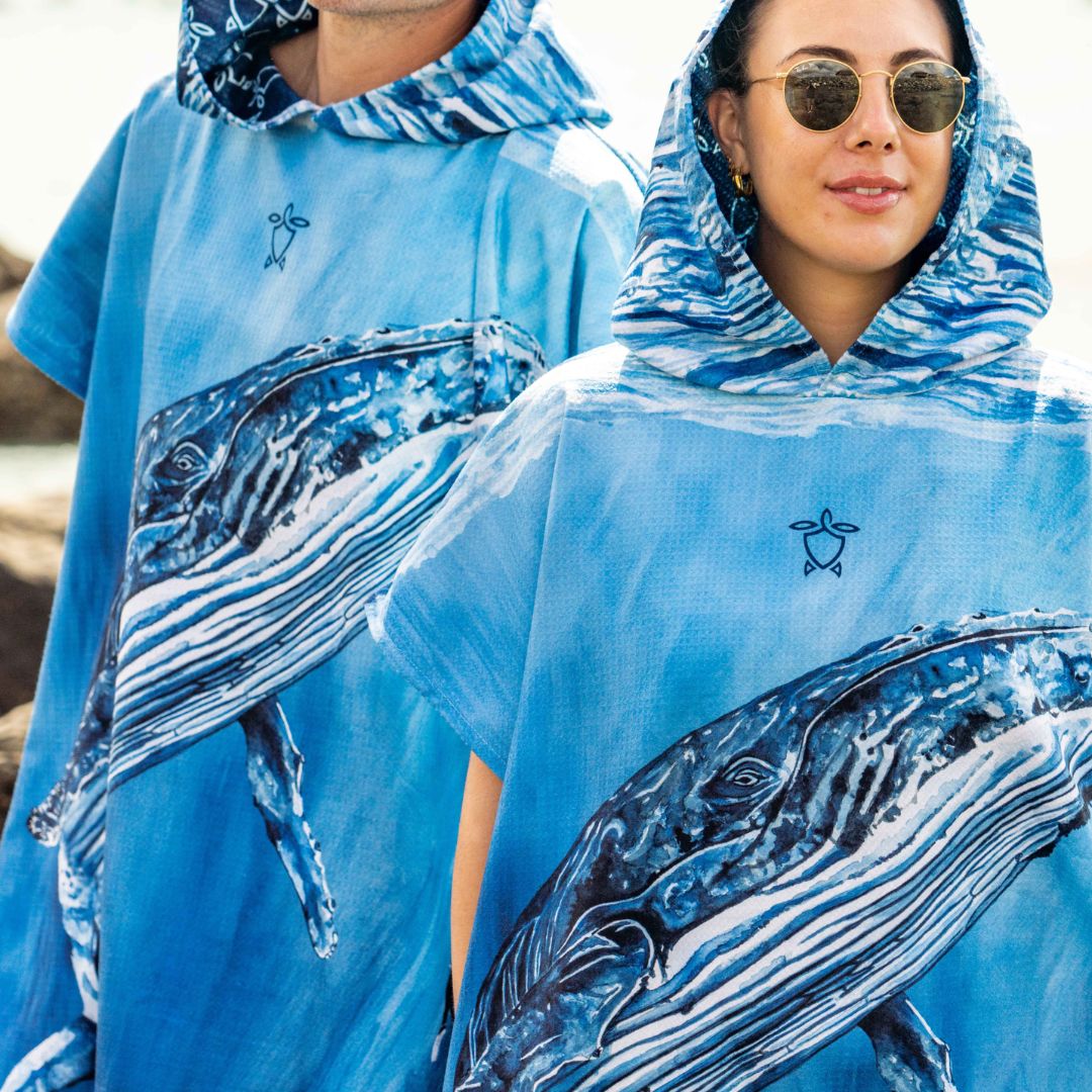 A close-up lifestyle shot of a woman wearing a light blue hooded poncho with a large blue and white whale print, along with a small turtle logo on the chest. A man is partially visible behind her wearing the same garment.