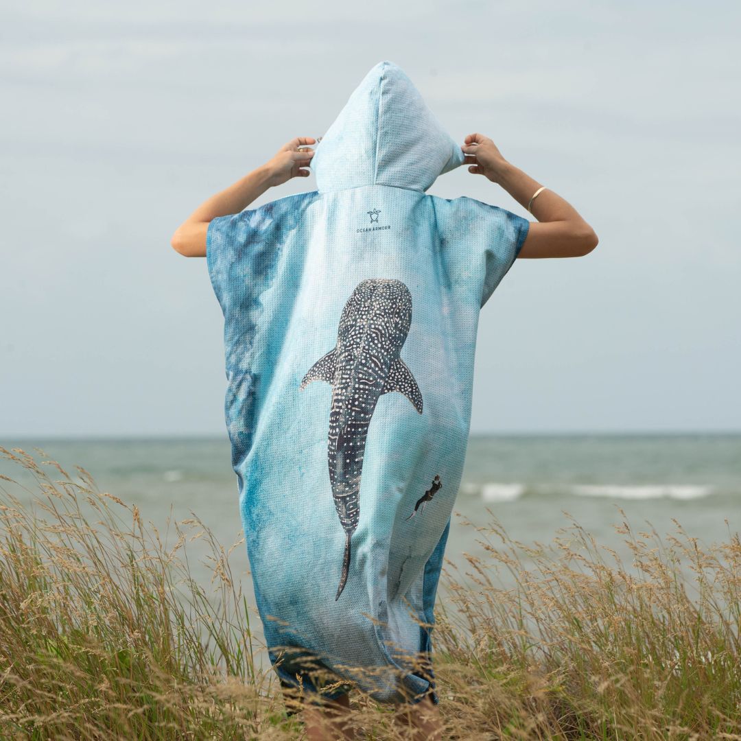 Back view of a person wearing a hooded Whale Shark Changing Poncho Towel while standing on coastal grass overlooking the ocean.