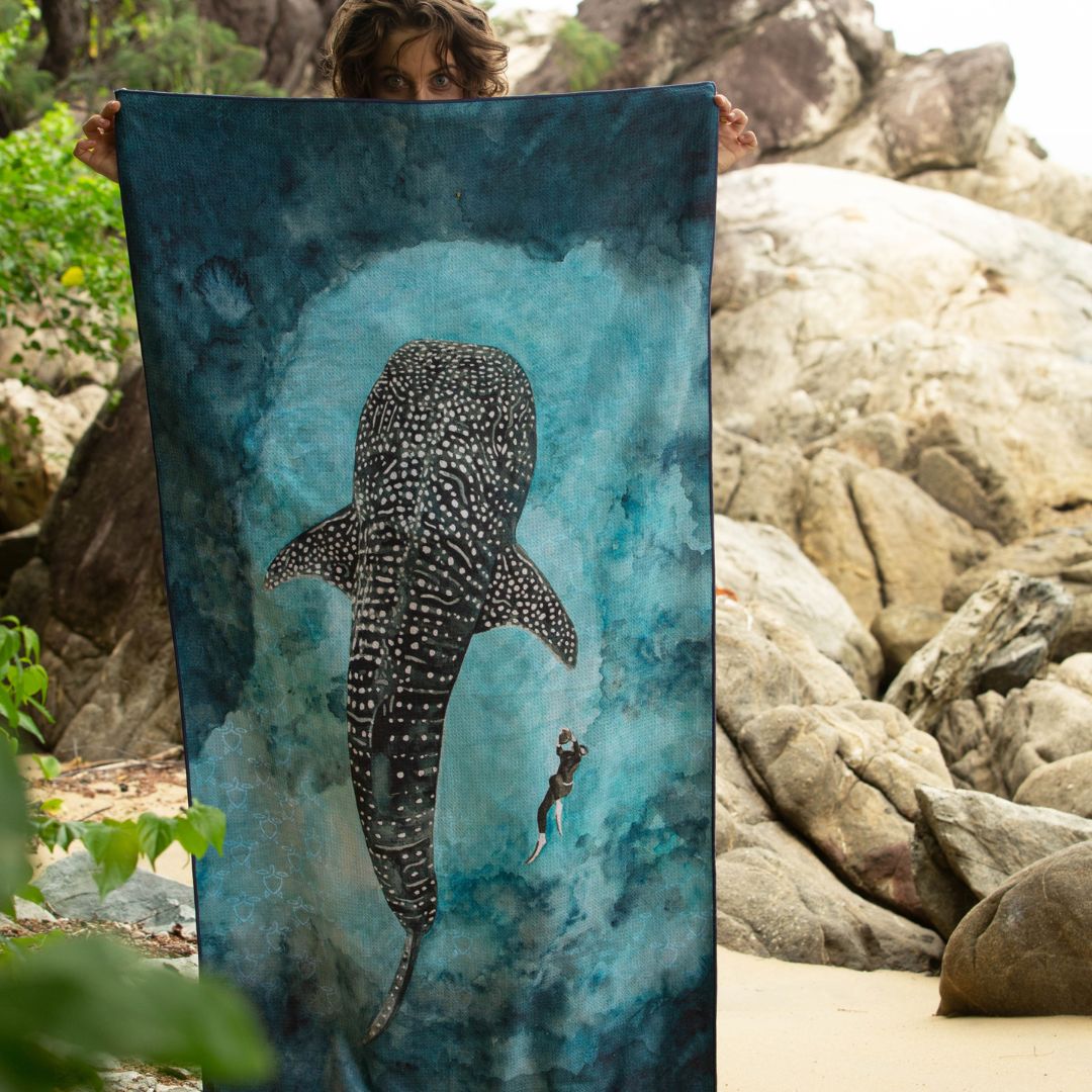 A person wearing a hooded beach towel or poncho with a watercolor-style manta ray and diver illustration in shades of blue and white, shown from the back against a light gray studio background.