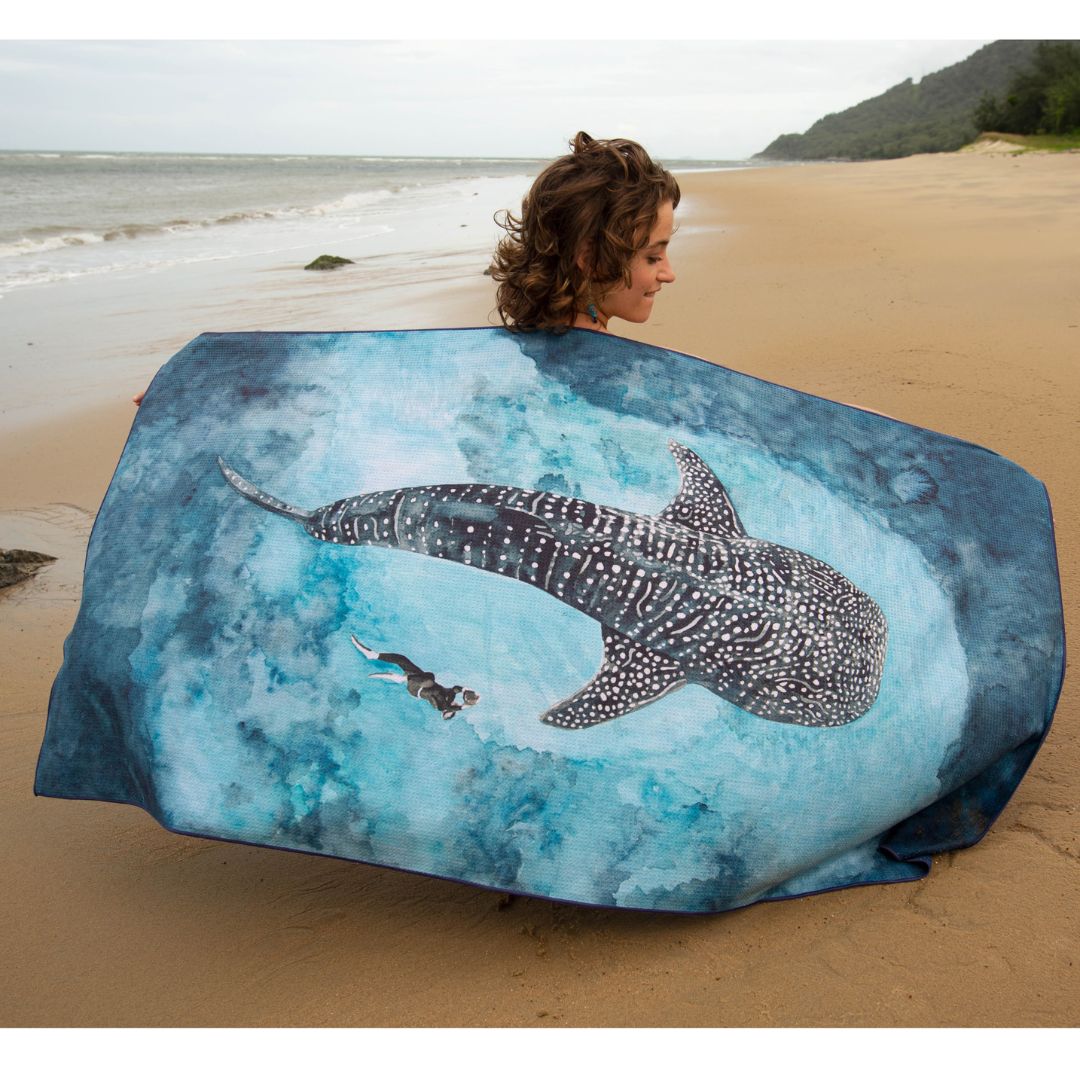 A woman on a sandy beach holds up a large printed towel or wrap featuring a whale shark and blue ocean water. The beach, ocean, and distant hillside are visible in the background.