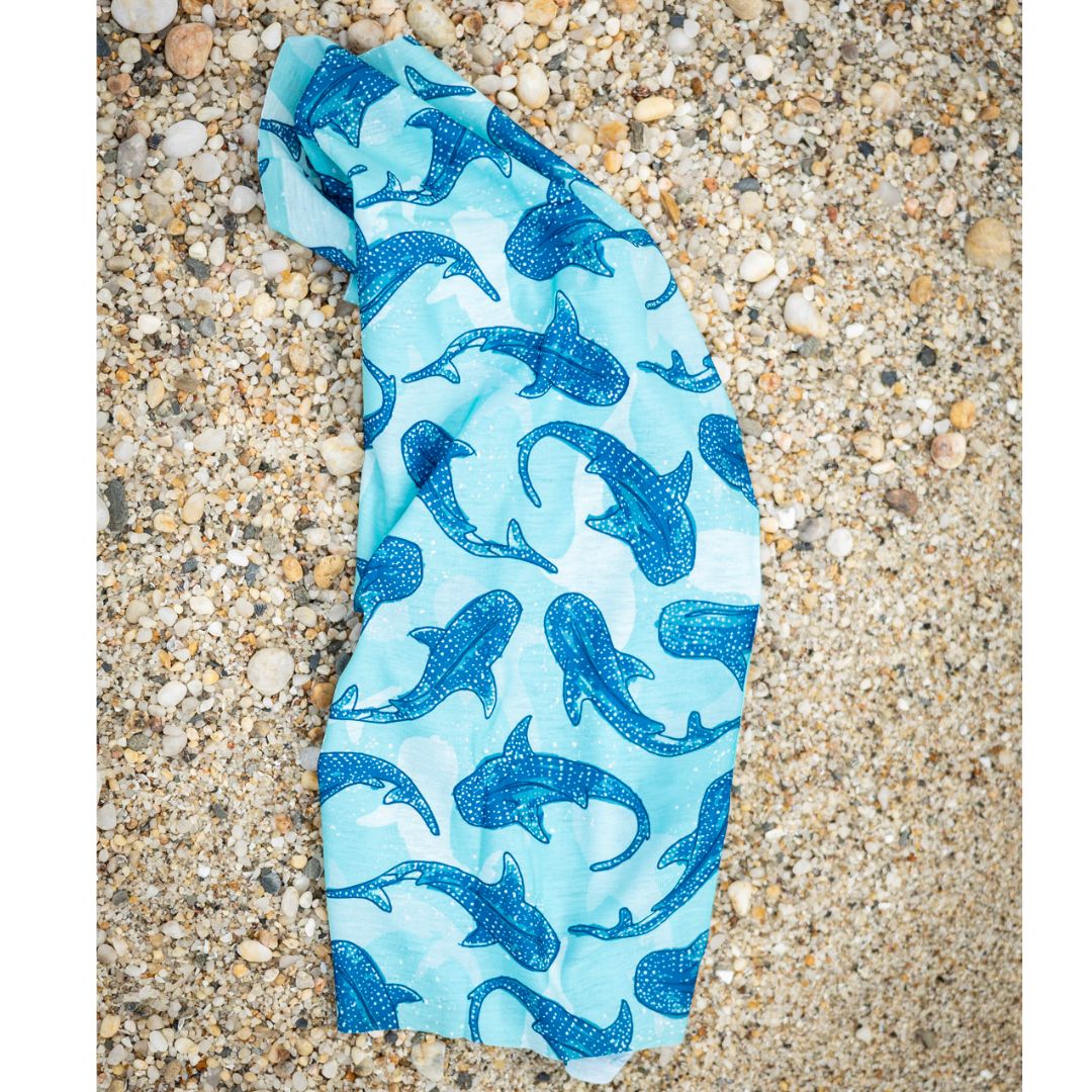 Whale Shark Sun Buff with a blue whale shark design on a light blue background, shown flat on a pebble beach