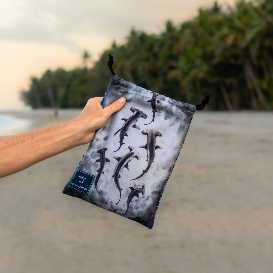 Person holding the carry pouch for the Hammerhead Shark quick-dry sand-free beach towel, featuring a watercolor-style hammerhead shark design on a gray ocean-inspired background.