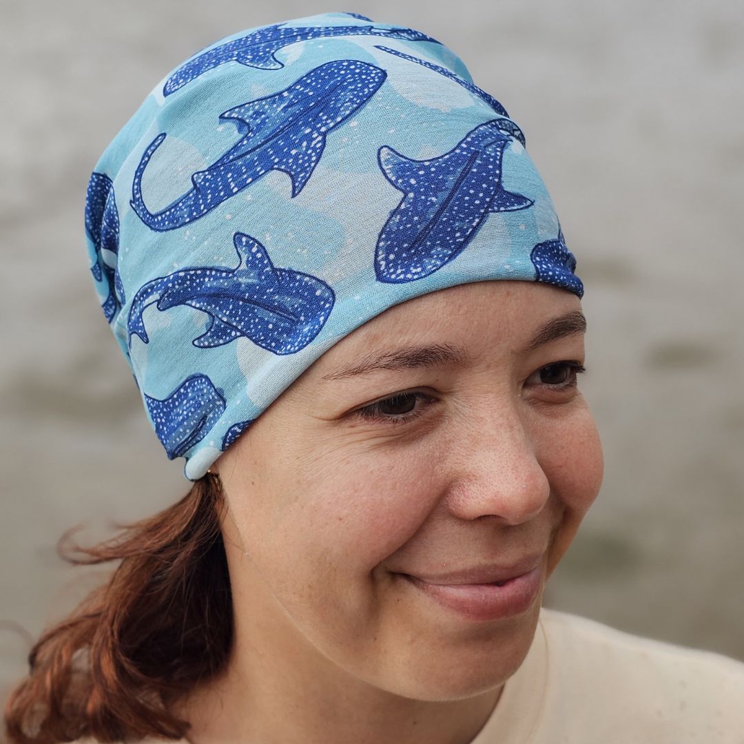A woman wearing a light blue swim cap with a blue whale shark pattern, photographed outdoors against a soft-focus gray background.