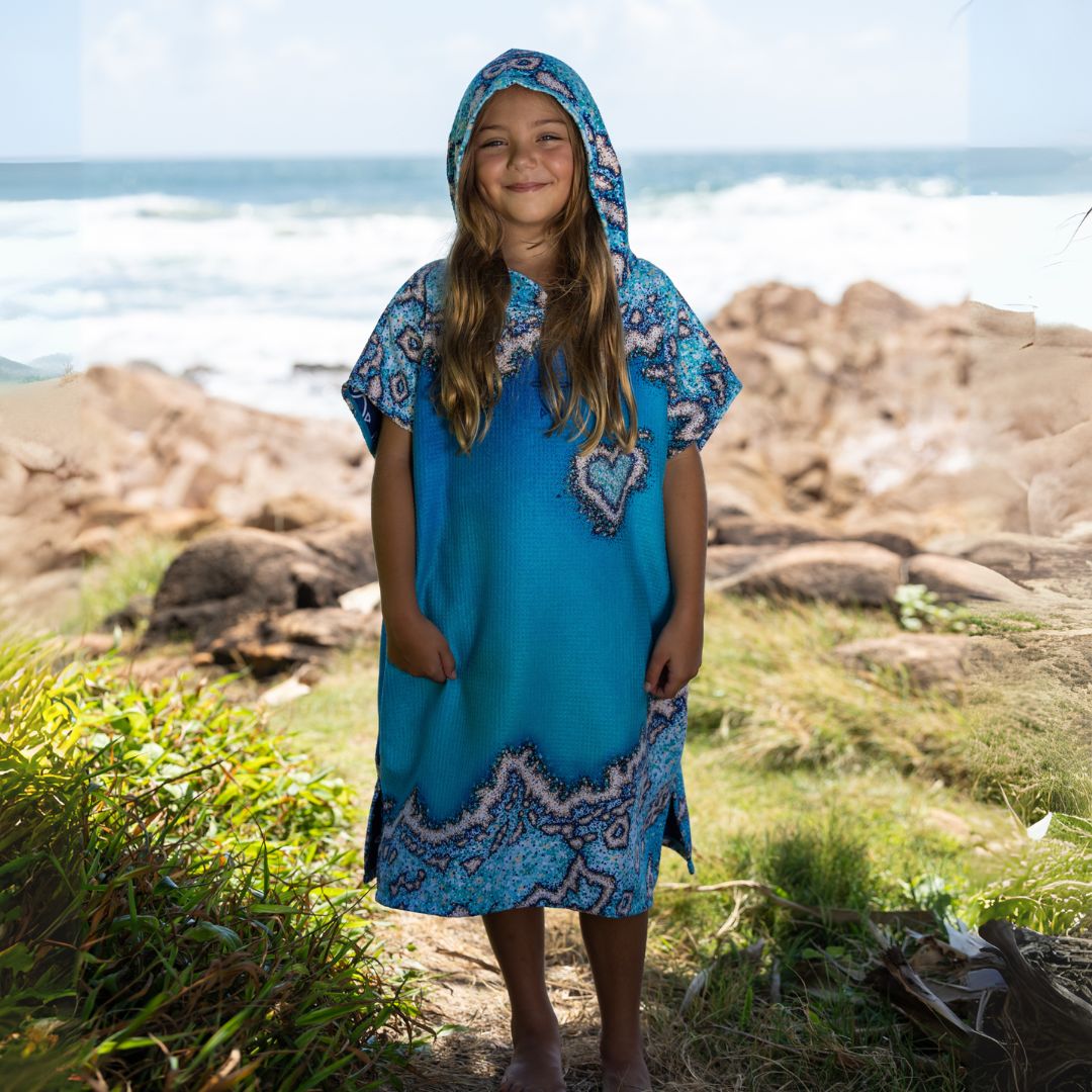 A young girl stands on a rocky beach wearing a bright turquoise blue hooded tunic dress with a white and dark blue paisley pattern. The ocean and rocks are visible in the background.