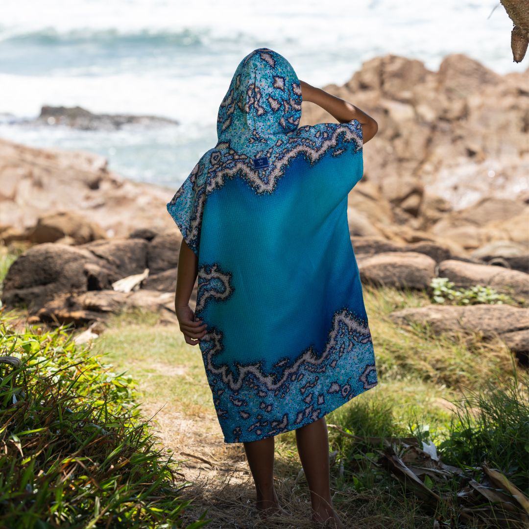 A kid standing on a rocky beach path wearing a bright blue hooded poncho with a paisley-style border pattern, with ocean and rocks in the background.