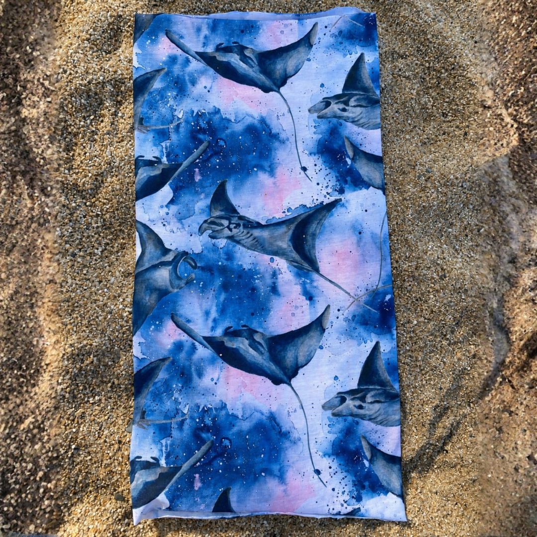 Manta Ray Multi-Use Sun Buff with a blue and purple watercolor ocean design, displayed flat on a sandy beach