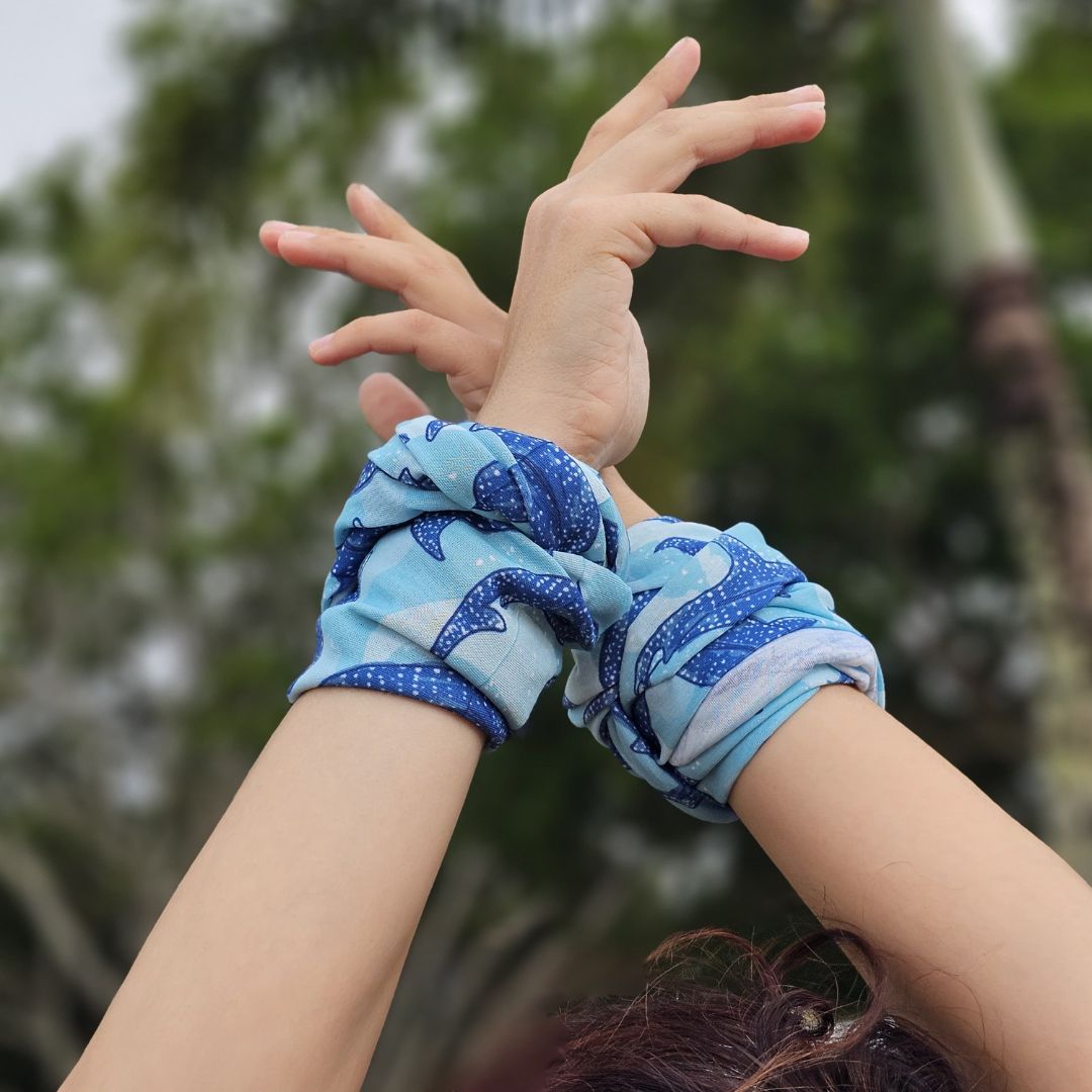 A close-up of a person's raised arms wearing two matching blue scrunchies with a shark print, set against a blurred outdoor background of green foliage.
