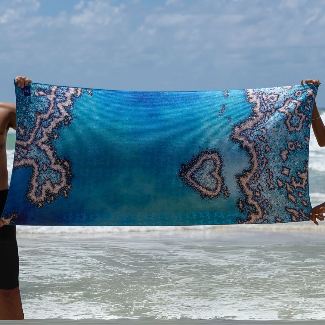 A person holds up a large rectangular beach towel with a blue ocean and coral reef pattern, with the ocean and sky visible in the background.