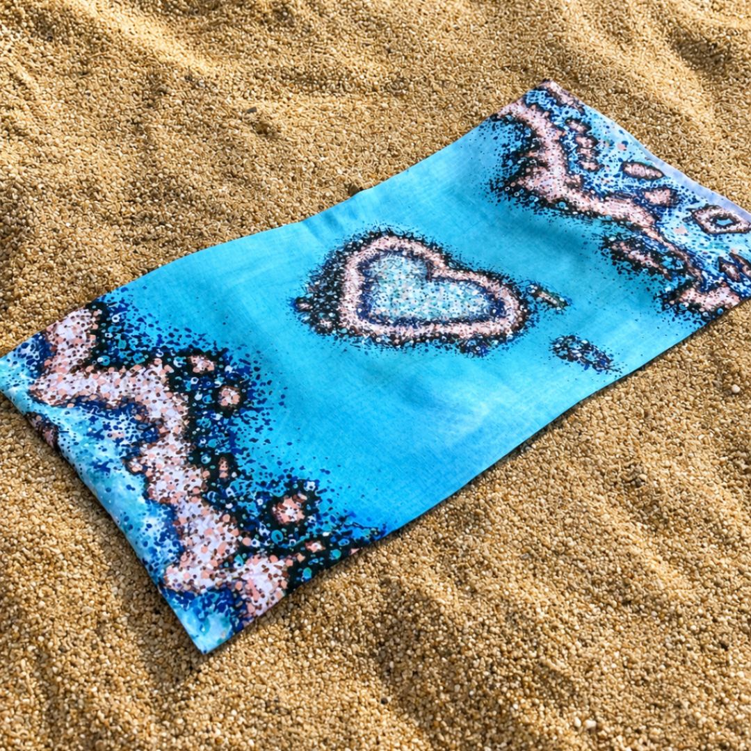 Blue Sun Buff featuring a heart-shaped coral reef illustration and coral accents around the edges, laid flat on beach sand.